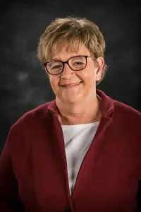 A woman with glasses and red jacket smiling.