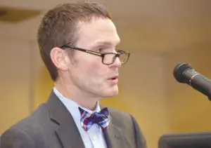 A man in glasses and a suit is speaking into a microphone.